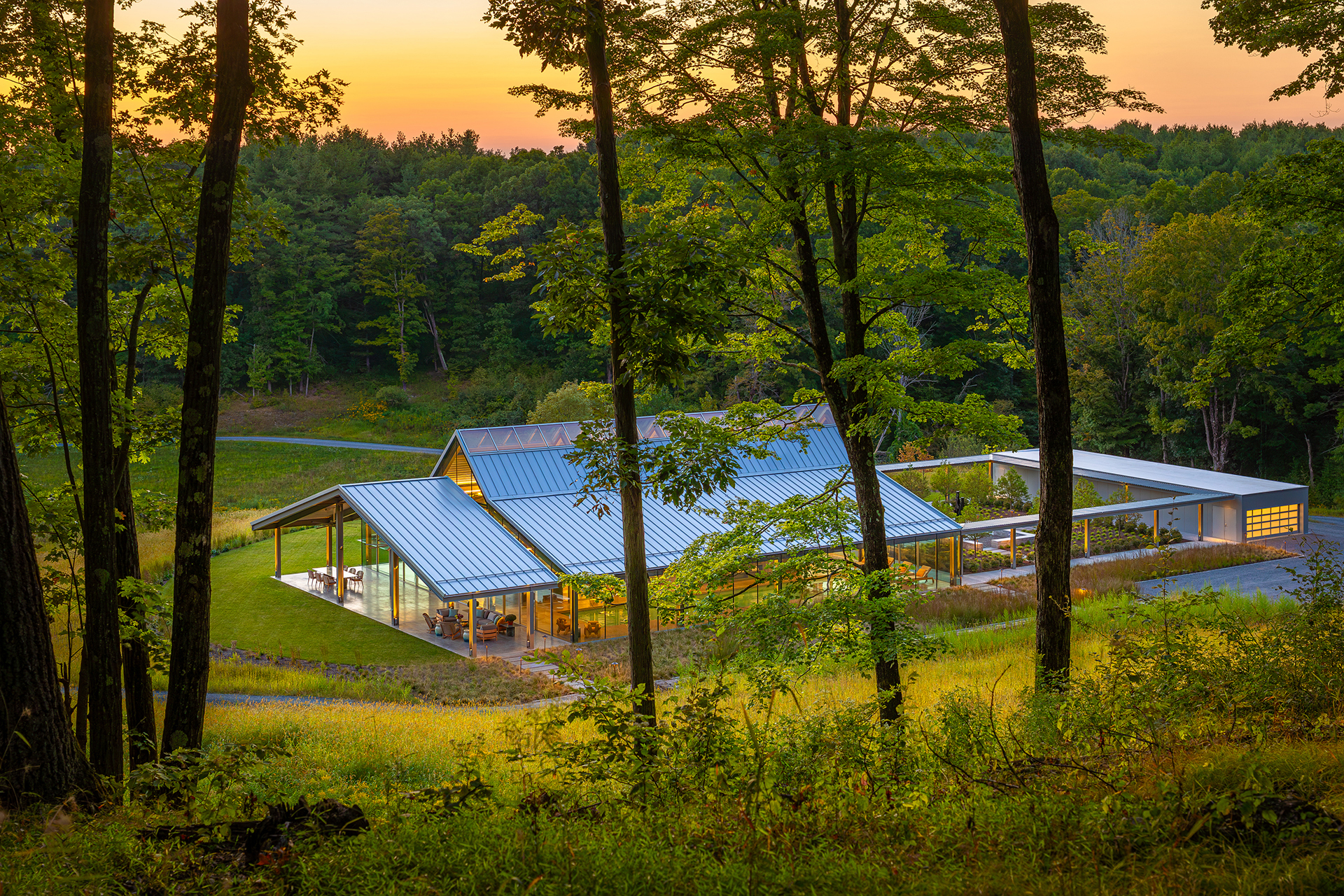 The pavilion's pitched metal roofs glowing at sunset, seen through the forest. Residential Recreation Pavilion, NY